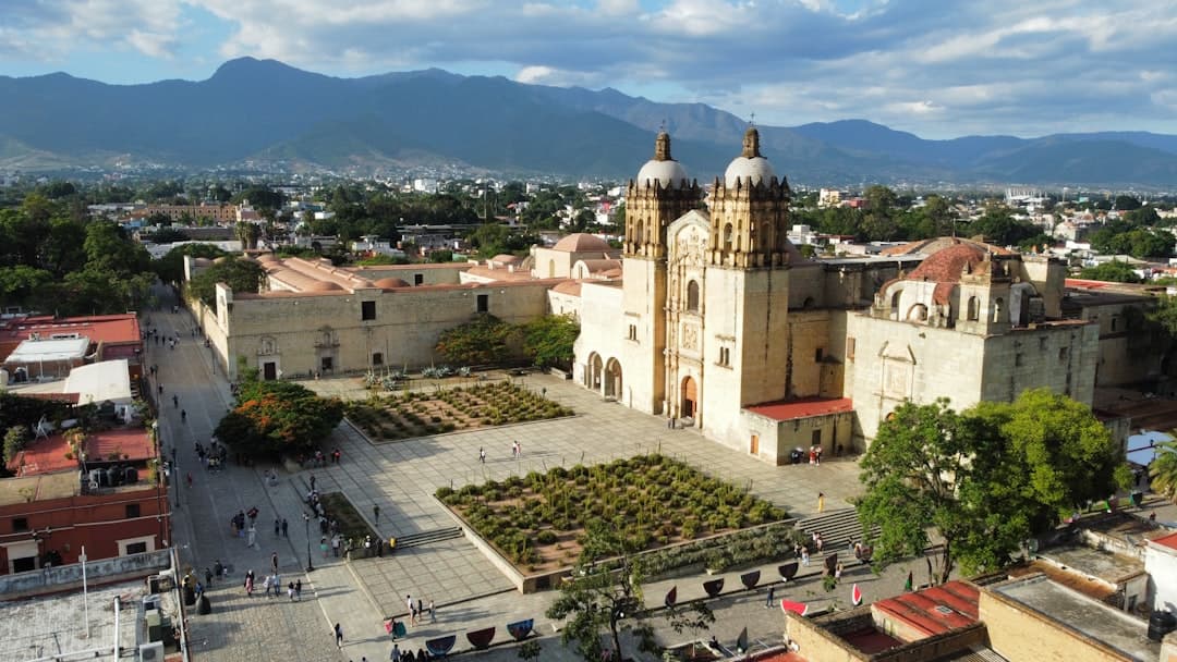 Aerial view of Oaxaca's historic city center featuring the Cathedral of Santo Domingo de Guzmán with its distinctive Baroque twin towers, surrounded by colonial architecture, manicured plaza grounds, and mountains in the background. - Photo by ryan doyle on Unsplash