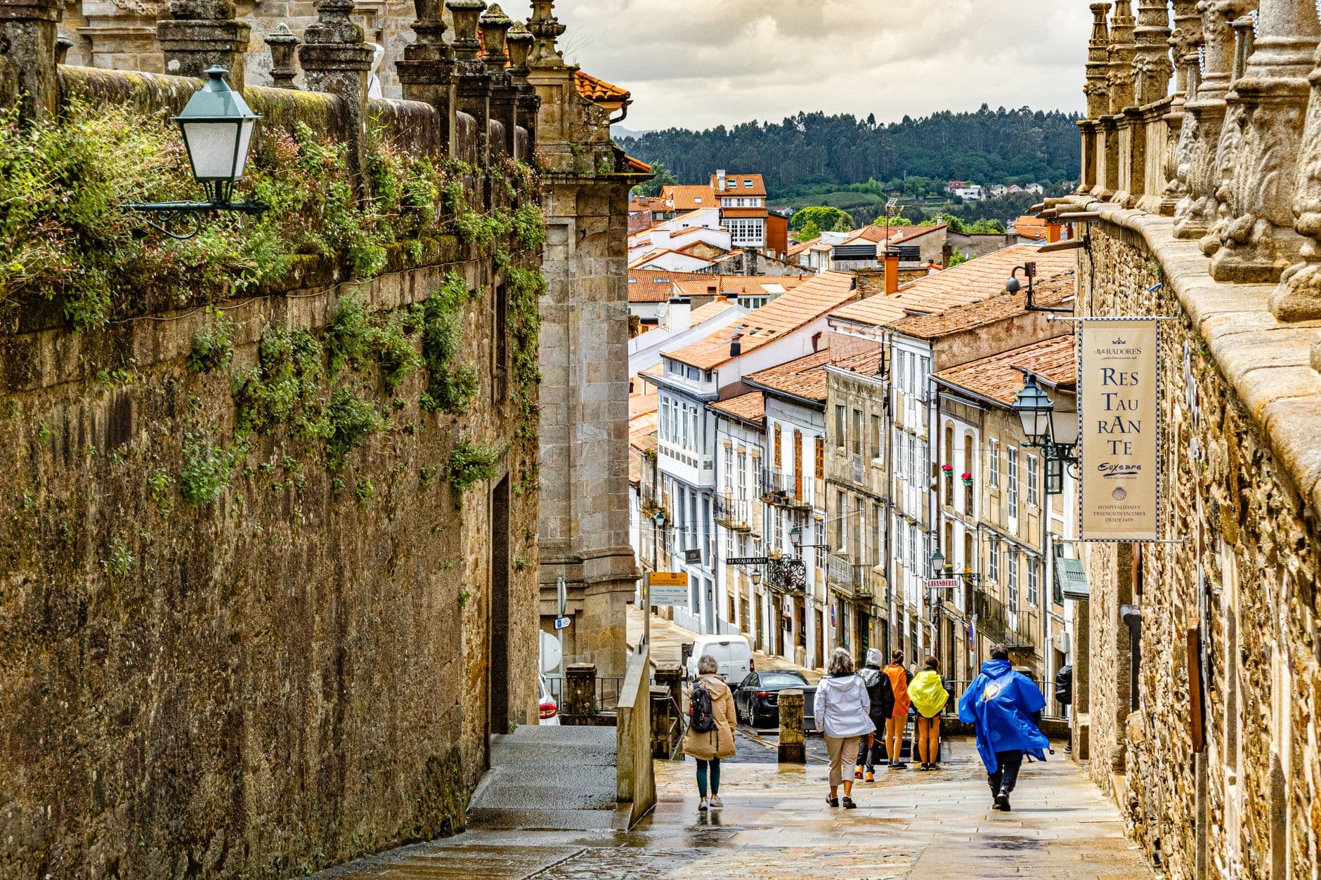 A narrow medieval stone street in Santiago de Compostela's old town descending toward the city center, with tourists walking along the wet cobblestones, flanked by granite buildings and traditional architecture, with the surrounding Galician landscape visible in the distance. - Photo by Jose Rodriguez Ortega on Pexels