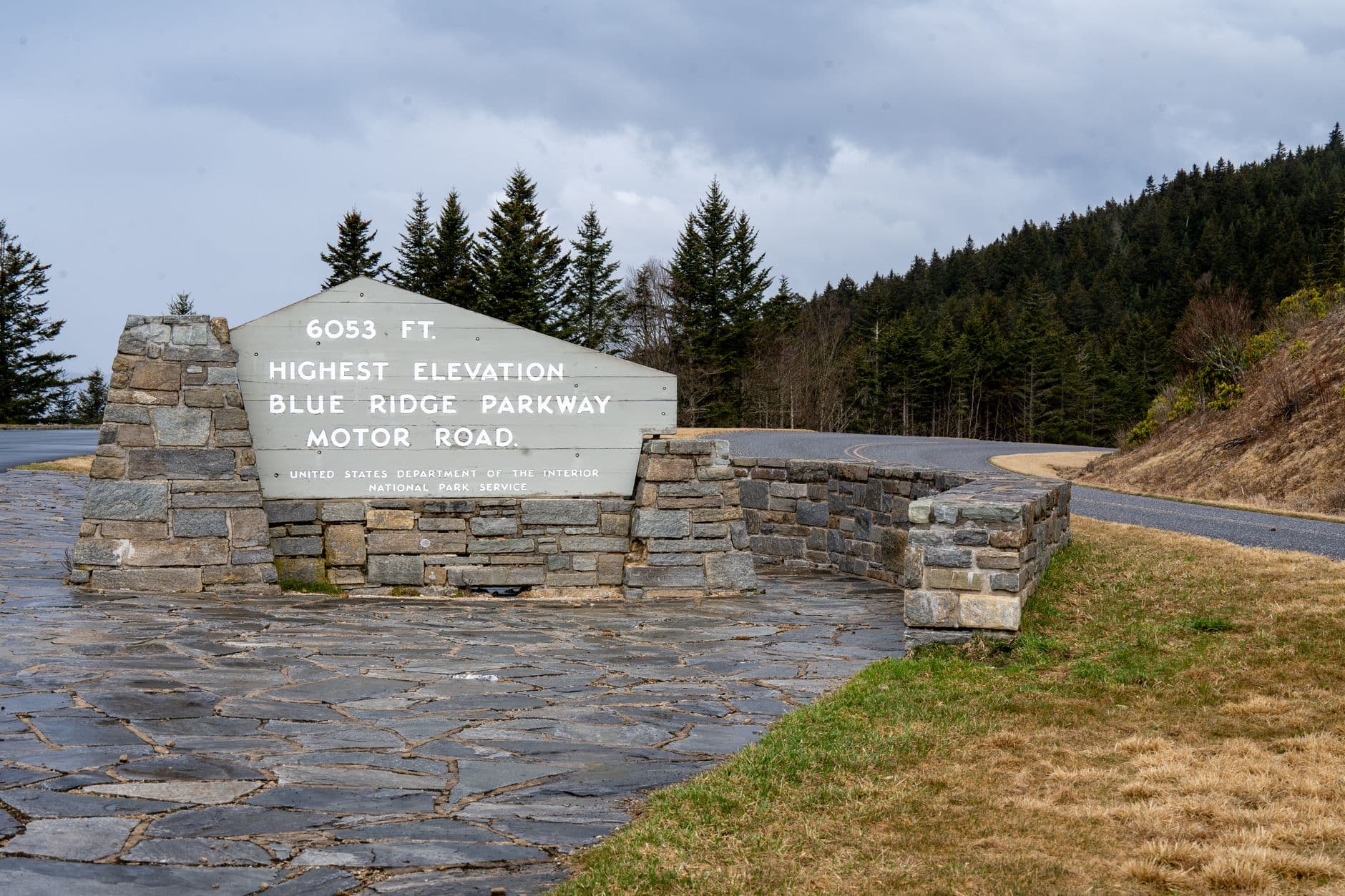 A stone marker at the highest point of the Blue Ridge Parkway Motor Road, situated along the roadside with coniferous forest and the parkway visible in the background. - Photo by Beth Fitzpatrick on Pexels