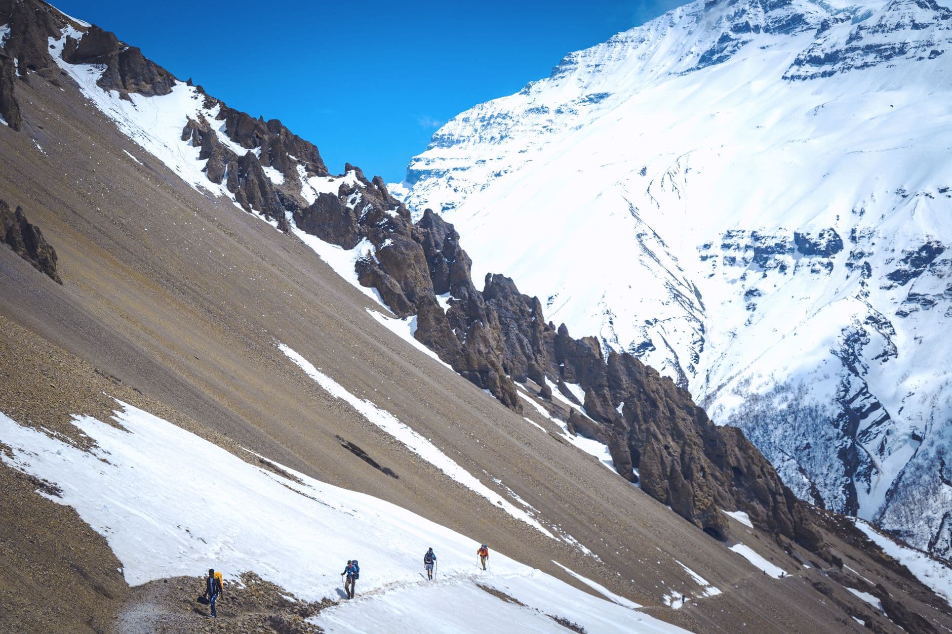 Trekkers ascending a high-altitude scree and snow slope in the Himalayas, with dramatic rocky ridgelines and snow-capped peaks visible in a mountain valley. - Photo by Ashok J Kshetri on Pexels