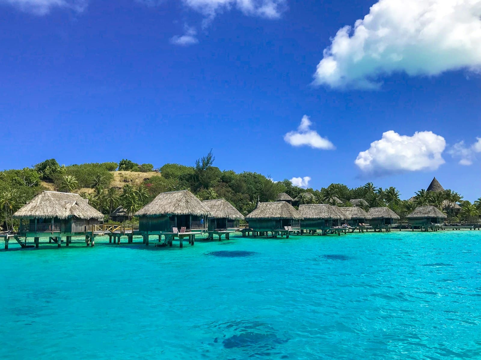 A row of overwater bungalows with thatched roofs on wooden stilts in crystalline turquoise lagoon water, with palm-covered hills behind and clear blue sky above. - Photo by Jo Barnes on Unsplash