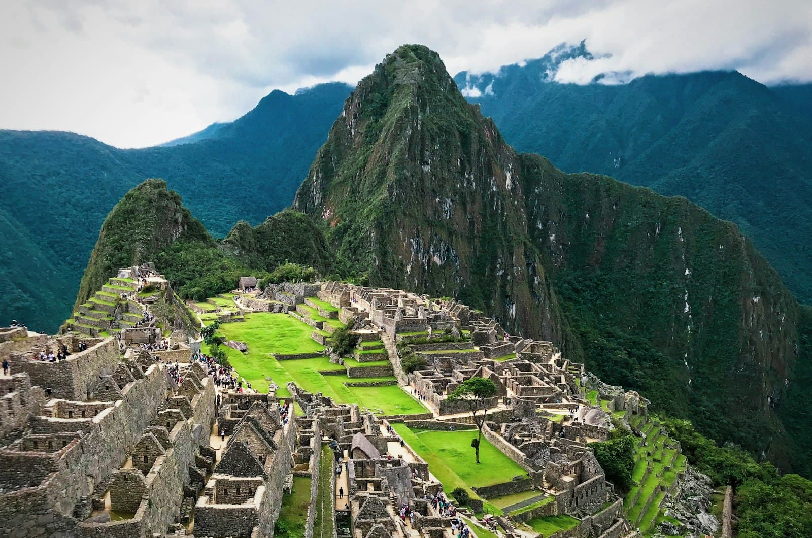 Machu Picchu, the 15th-century Incan citadel, photographed from an elevated vantage point showing the central plaza, surrounding stone structures, and Huayna Picchu peak rising behind, with forested Andean mountains in the misty background. - Photo by Eddie Kiszka on Unsplash