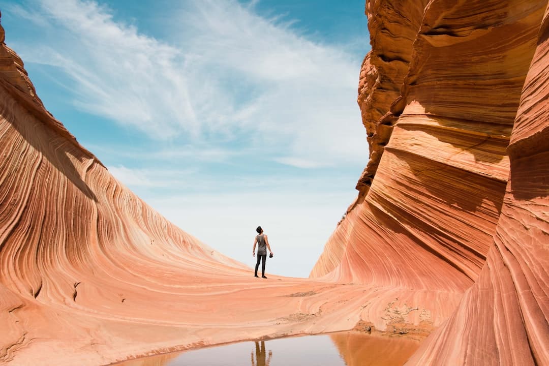 A solitary hiker stands in a narrow slot canyon with dramatically striated Navajo Sandstone walls on either side, with a small pool of water reflecting the canyon floor below a blue sky. - Photo by Christopher Ruel on Unsplash