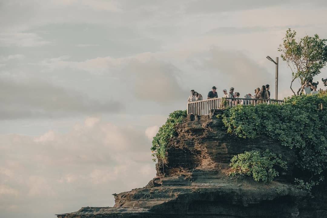 Tourists gathered on a modern wooden railing platform built into a dramatic cliffside overlooking clouds and ocean, with tropical vegetation and modern infrastructure visible. - Photo by Dennis Schmidt on Unsplash