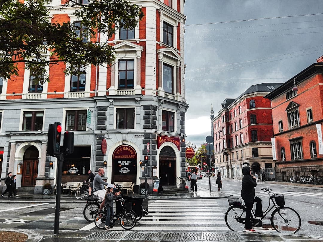An urban street scene in Copenhagen showing red-brick period buildings, pedestrians, cyclists on a paved plaza with a crosswalk, and characteristic Danish urban infrastructure. - Photo by flickch on Unsplash