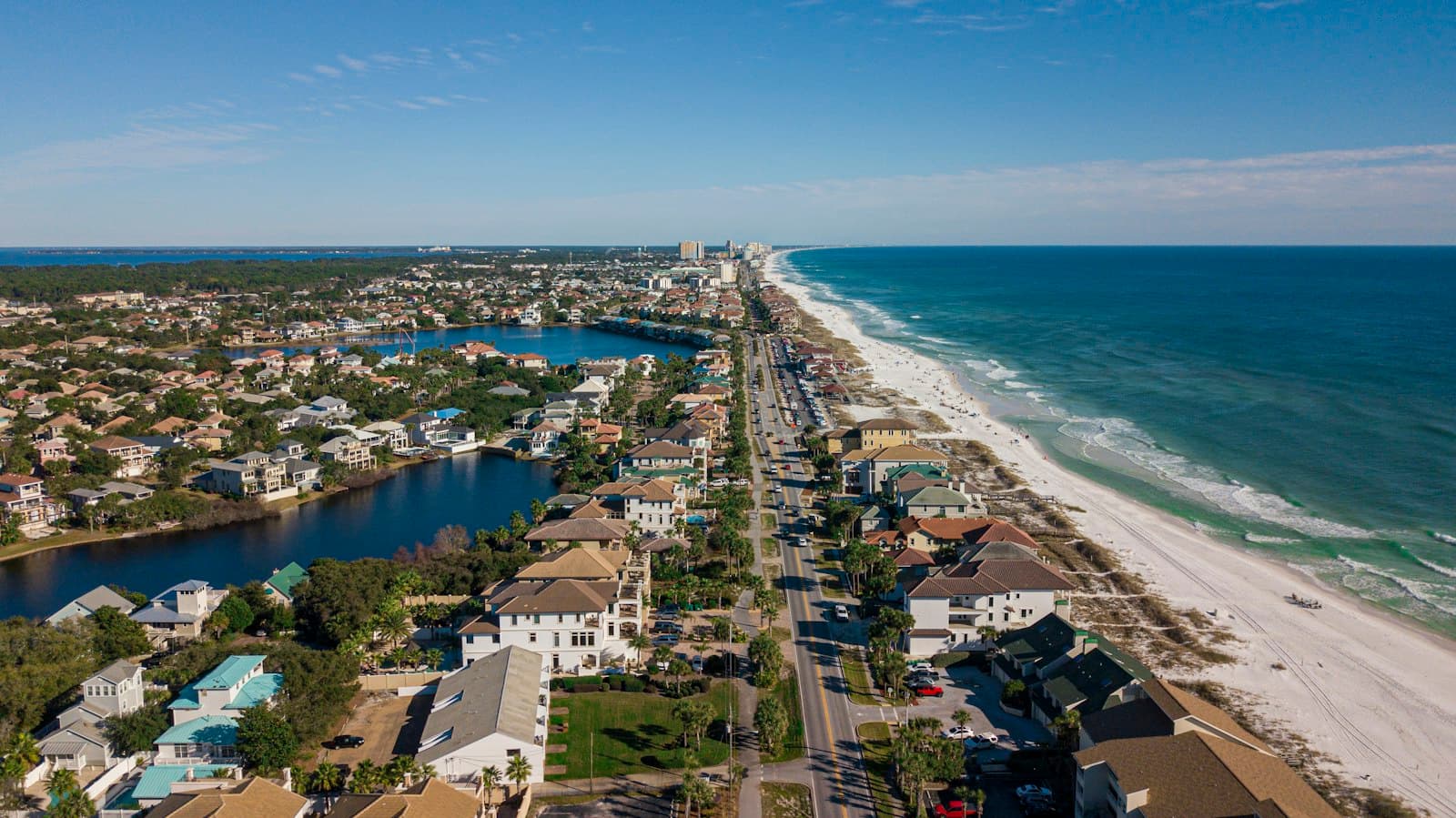 Aerial view of a beachfront coastal town with high-rise hotels and condos along the beach, residential neighborhoods with pools and waterfront properties inland, a main coastal road running parallel to a white sand beach and emerald Gulf waters, and inland waterways and lakes characteristic of a barrier island community. - Photo by TopSphere Media on Unsplash