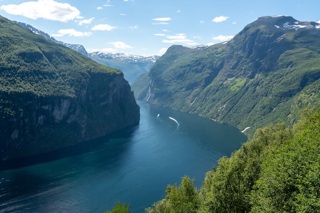 A sweeping aerial view of a deep Norwegian fjord with steep forested mountainsides, a visible waterfall, and a small cruise ship navigating the turquoise waters, with snow-patched peaks visible in the distance. - Photo by Sven on Unsplash