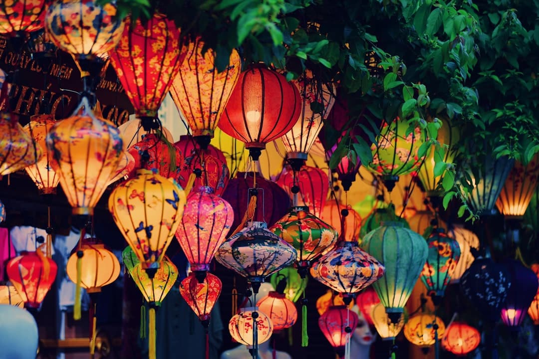 Illuminated traditional silk lanterns in vibrant reds, yellows, and oranges hanging above a street at night, with ivy and foliage visible on the right side. - Photo by Hieu Do Quang on Unsplash