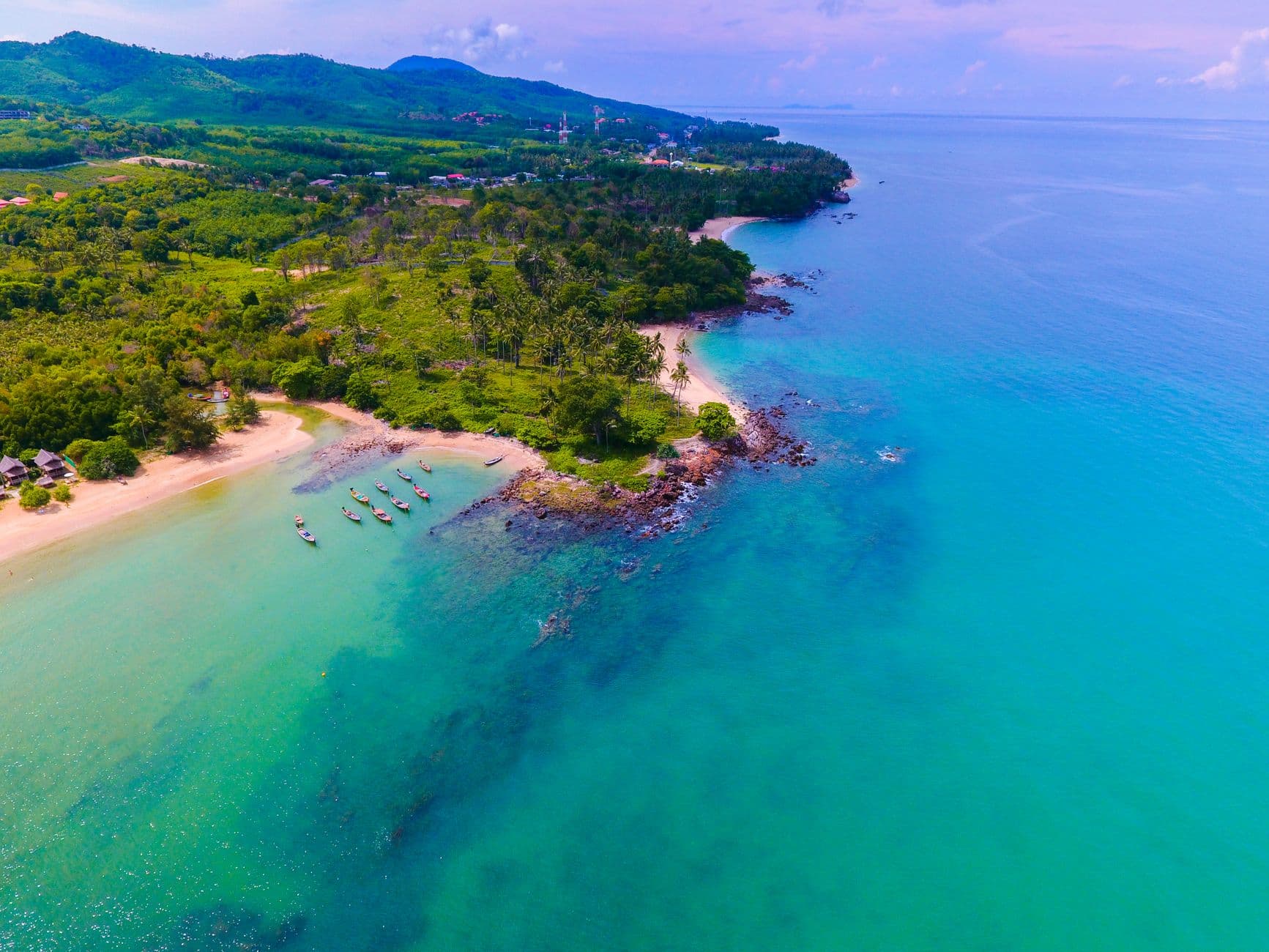 An aerial view of a pristine tropical beach with turquoise waters, pink sand, wooden fishing boats, dense jungle vegetation, and mountainous backdrop characteristic of southern Thai islands. - Photo by Humphrey Muleba on Pexels