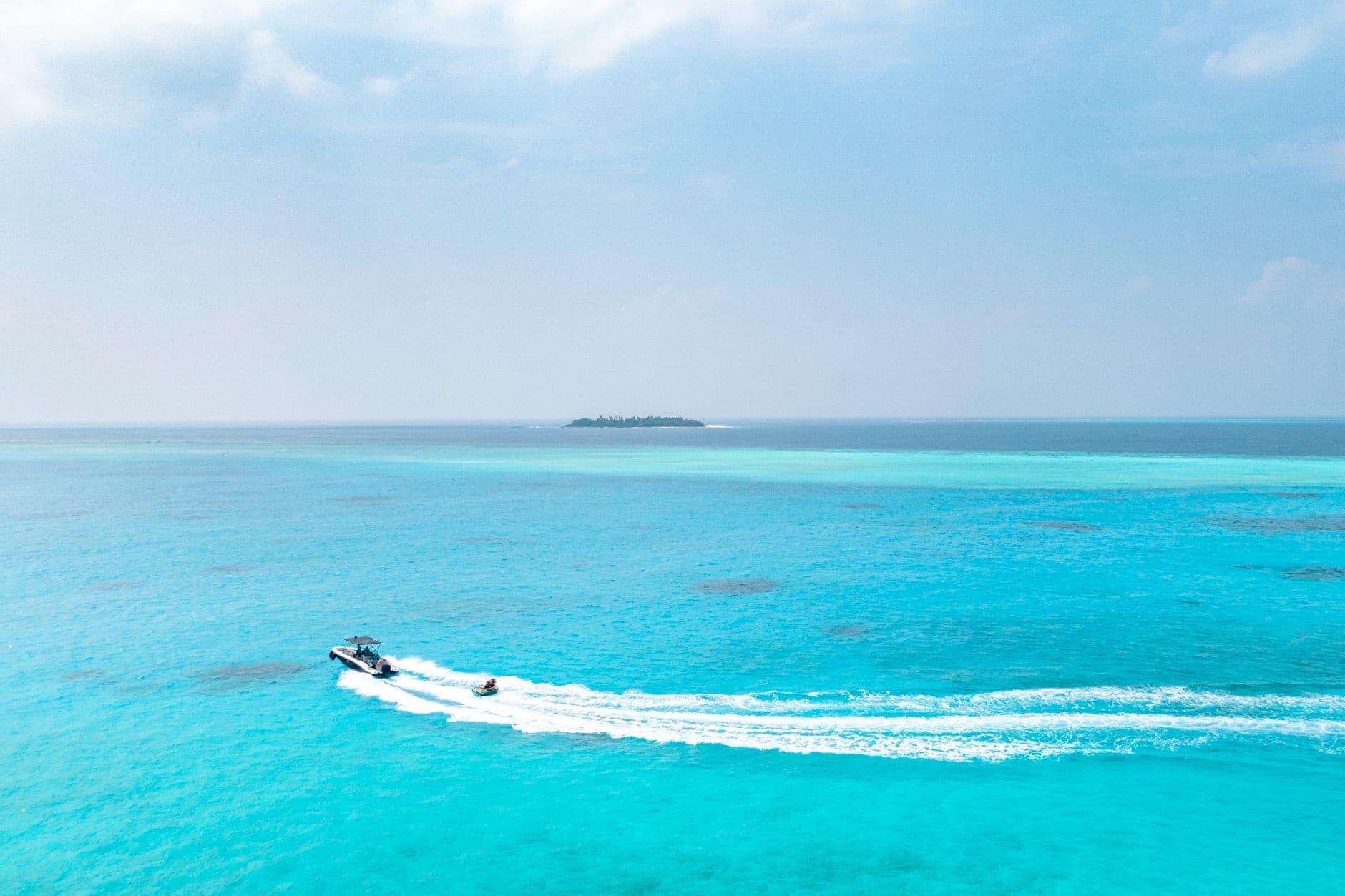 A speedboat creates a wake in the shallow turquoise waters of a tropical atoll lagoon, with a small island visible on the horizon and clear blue skies above. - Photo by Asad Photo Maldives on Pexels