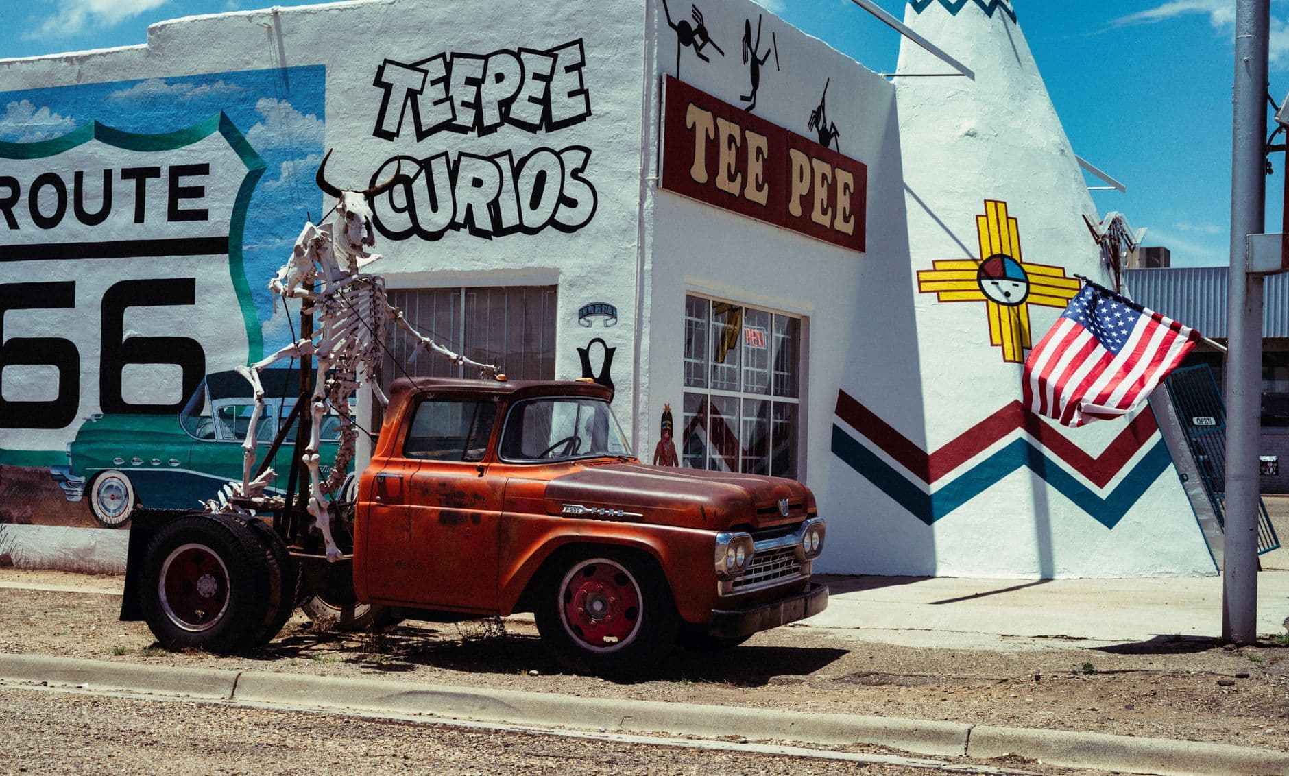 A vintage red Ford pickup truck with skeleton decorations parked in front of Route 66-themed roadside buildings, including a 'Teepee Curios' shop with Route 66 signage and New Mexico state symbols. - Photo by Alfo Medeiros on Pexels