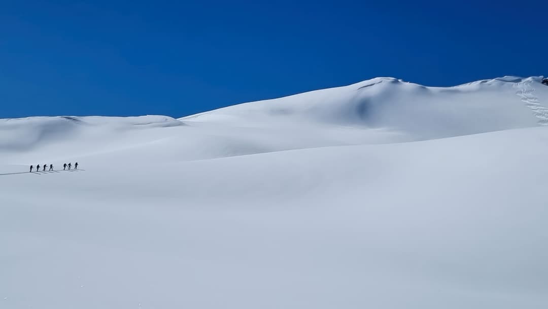 A group of backcountry skiers traverse a vast, snow-covered alpine slope under clear blue sky, with rolling snow-laden peaks visible in the background. - Photo by Hans-Jurgen Mager on Unsplash