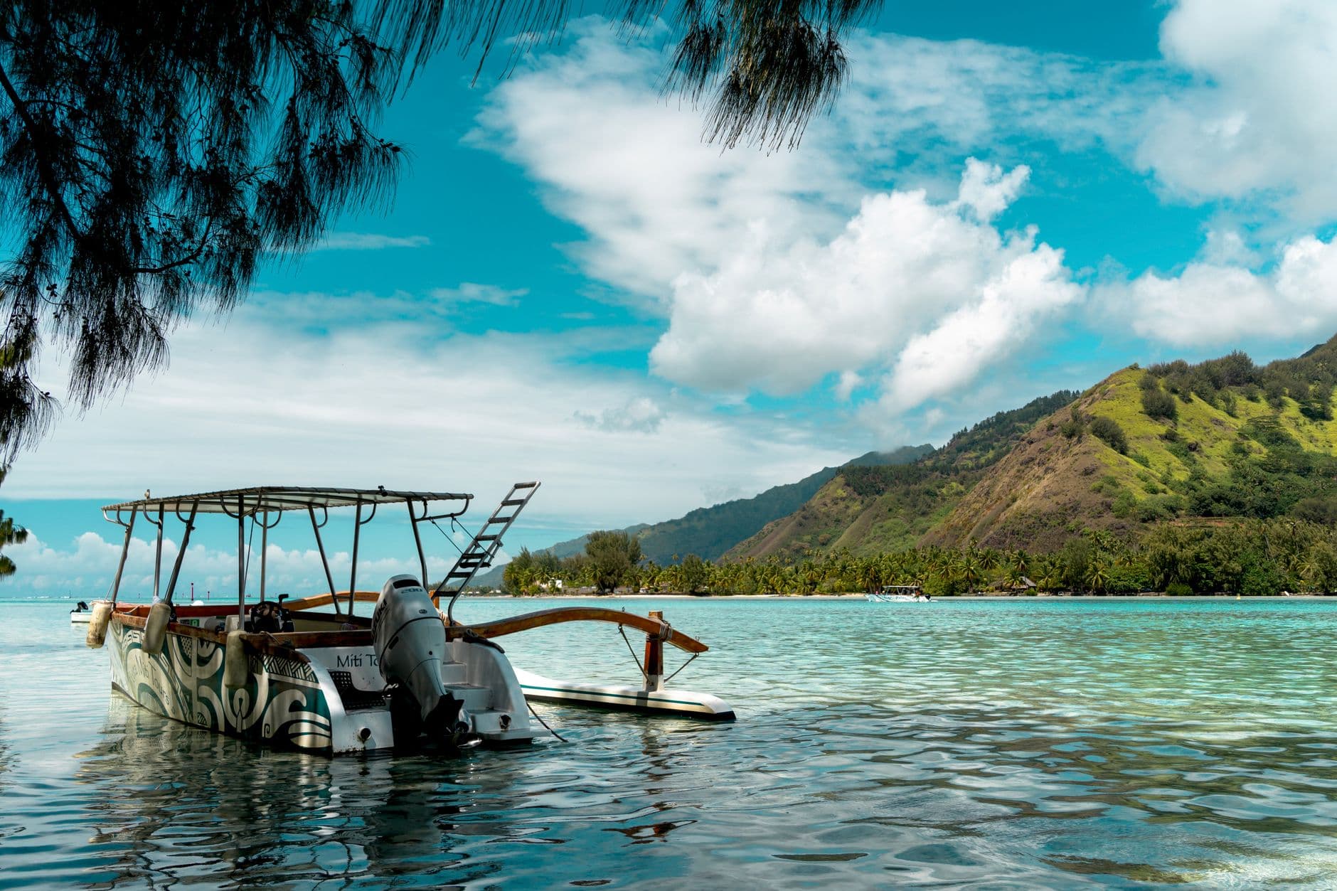 A traditional outrigger canoe with a black outboard motor and canopy sits in shallow turquoise lagoon water, with a steep green volcanic island and palm trees visible across the water. - Photo by Adrien Daurenjou on Pexels