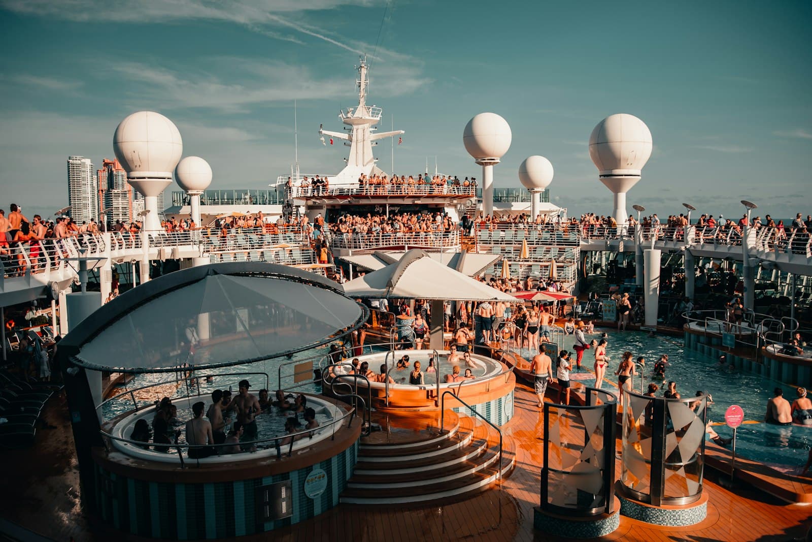 A crowded cruise ship's lido deck during sea day, with multiple pools, waterslides, sunbathing passengers, and the vessel's radar domes and superstructure visible under clear skies. - Photo by Colin Lloyd on Unsplash