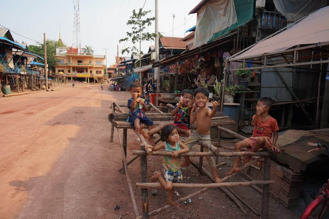 A village street scene in Cambodia showing local children playing on wooden benches, with traditional wooden stilt houses, shops with tarps, and a golden temple spire visible in the background. - Photo by Dylan Winterflood on Unsplash