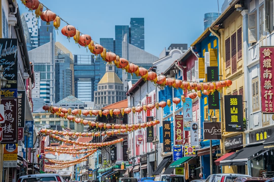A vibrant street in Singapore's Chinatown decorated with red lanterns strung overhead, lined with traditional colorful shop-houses and modern signage, with tall office buildings visible in the background under clear blue sky. - Photo by Adrian Jakob on Unsplash