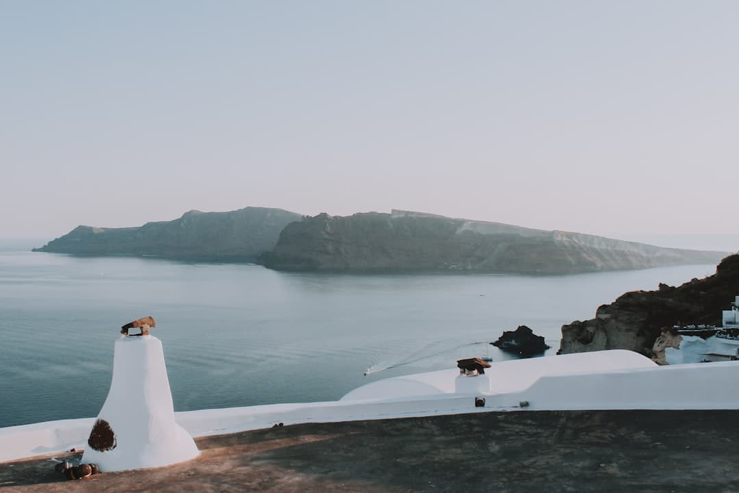 A white-washed cliffside structure with traditional Cycladic domed architecture overlooks the Santorini caldera, with the volcanic island visible across calm blue waters. - Photo by Nate Johnston on Unsplash