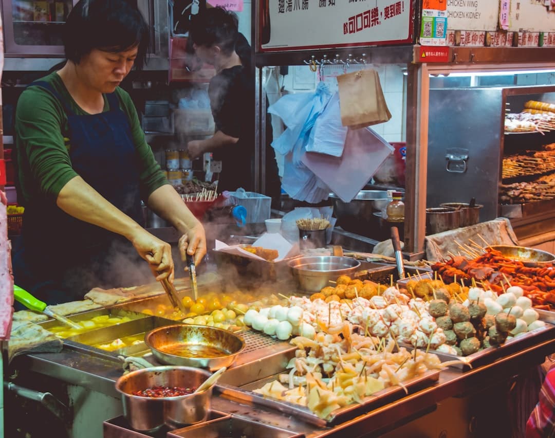 A street food vendor in a Chinese market stall prepares skewered food items and grilled meats, with various meat and vegetable skewers arranged on metal trays and sauce bowls visible. - Photo by Vernon Raineil Cenzon on Unsplash