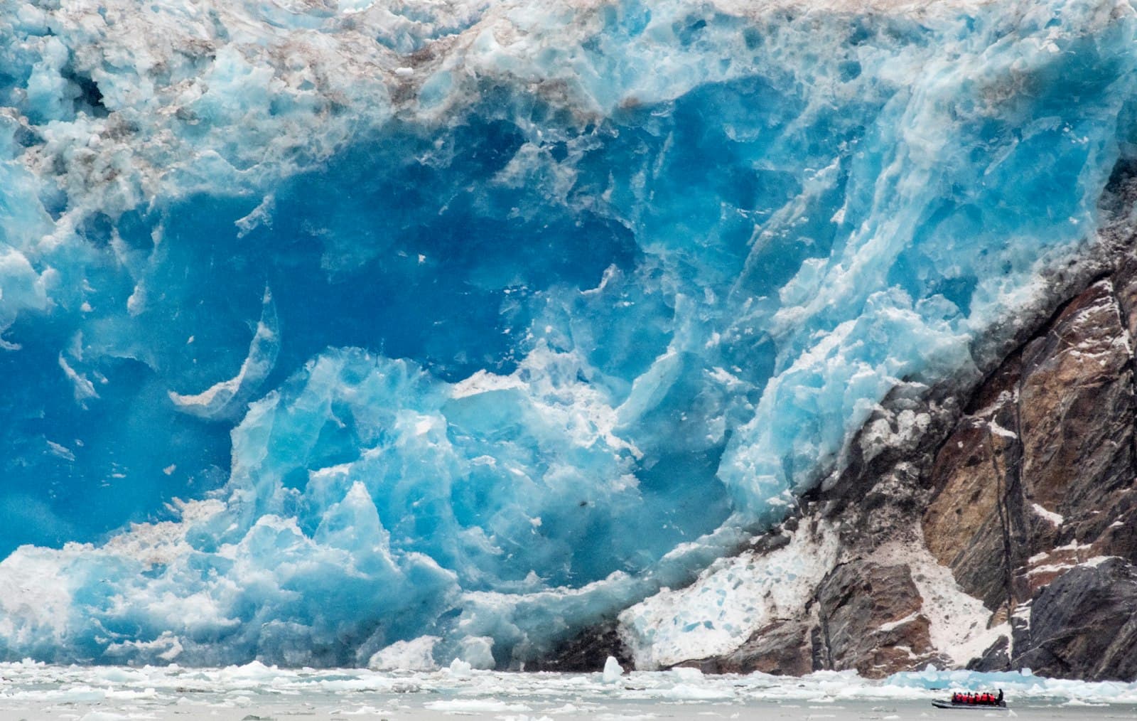 A tidewater glacier calves into the sea, with a small tour boat positioned in the foreground to show scale, demonstrating the dramatic ice formations and water activity typical of Alaska's Inside Passage. - Photo by John Yunker on Unsplash