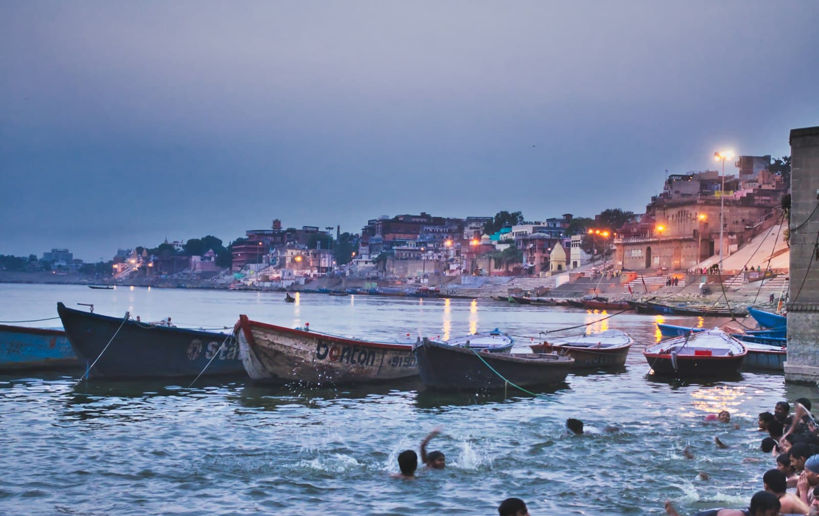 An evening scene of Varanasi's ghats along the Ganges River, showing multi-colored buildings lit by street lights, traditional wooden boats moored in the water, and people bathing and wading in the river. - Photo by Shiv Prasad on Unsplash