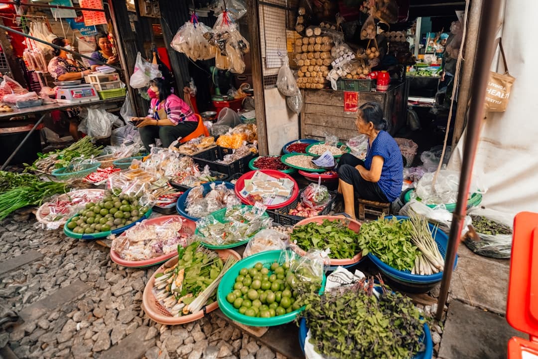 A bustling street market stall in what appears to be a Bangkok wet market, with vendors selling fresh vegetables, herbs, limes, and prepared foods displayed in colorful plastic bowls and basins. - Photo by Mauro Lima on Unsplash