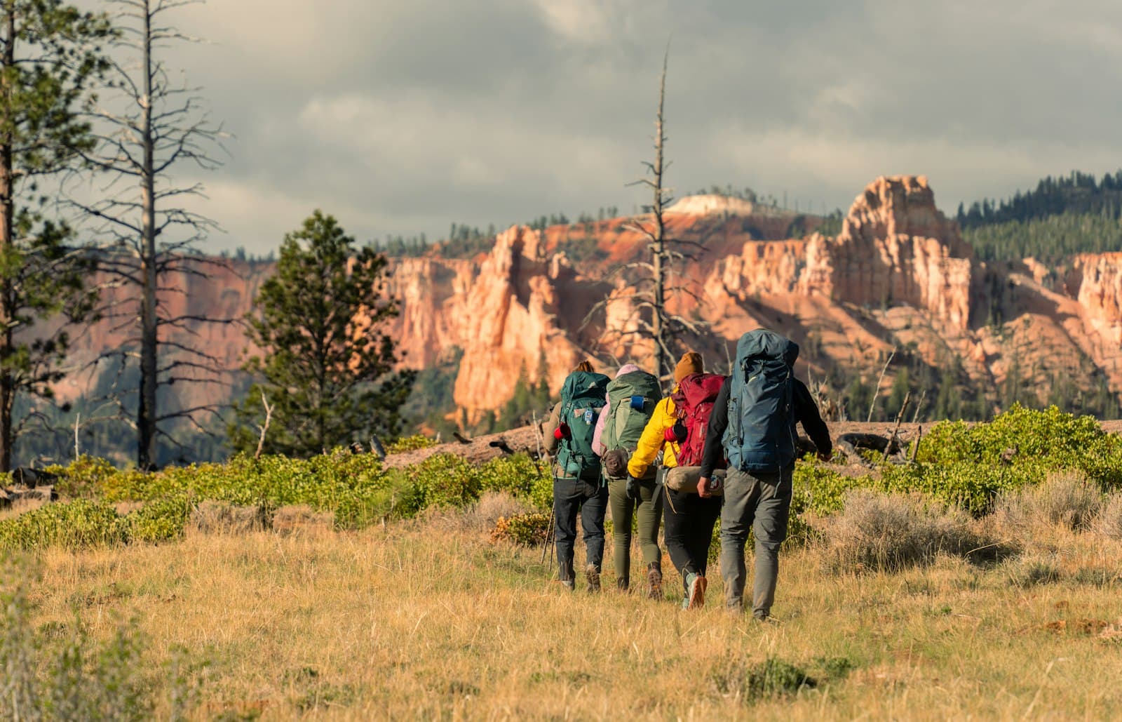 Five hikers with backpacks walking across sagebrush meadow toward distinctive hoodoo formations rising in the distance under partly cloudy skies. - Photo by Alex Moliski on Unsplash