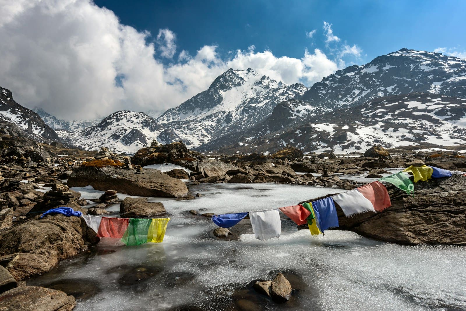 A high-altitude alpine stream flowing through rocky terrain with snow-capped Himalayan peaks in the background, adorned with colorful Buddhist prayer flags strung across rocks. - Photo by Sergey Pesterev on Unsplash
