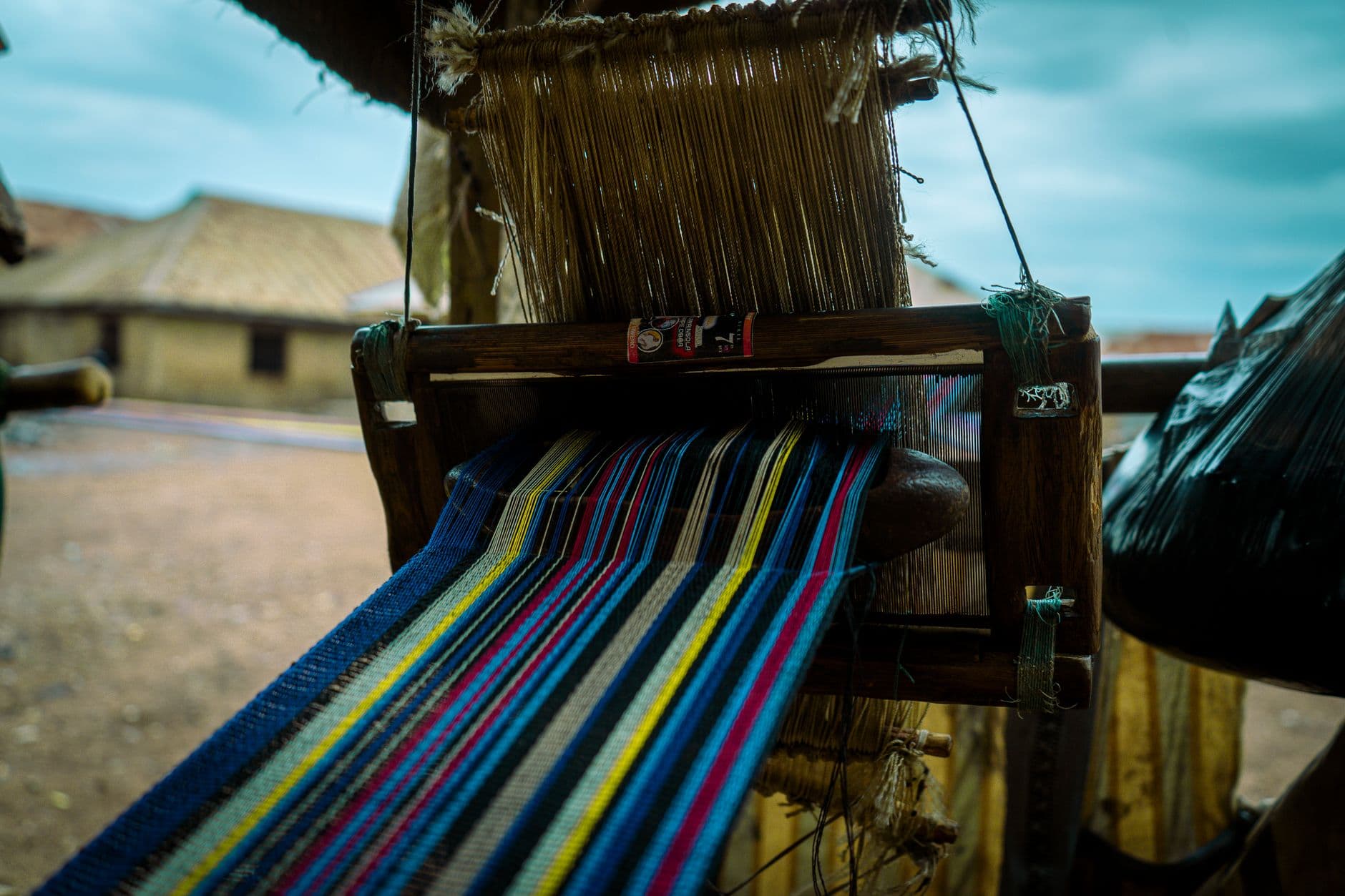 A close-up of a traditional Ashanti kente loom showing colorful striped thread being woven, with thatched-roof structures and sandy ground visible in the background. - Photo by Silvanus Solomon on Pexels