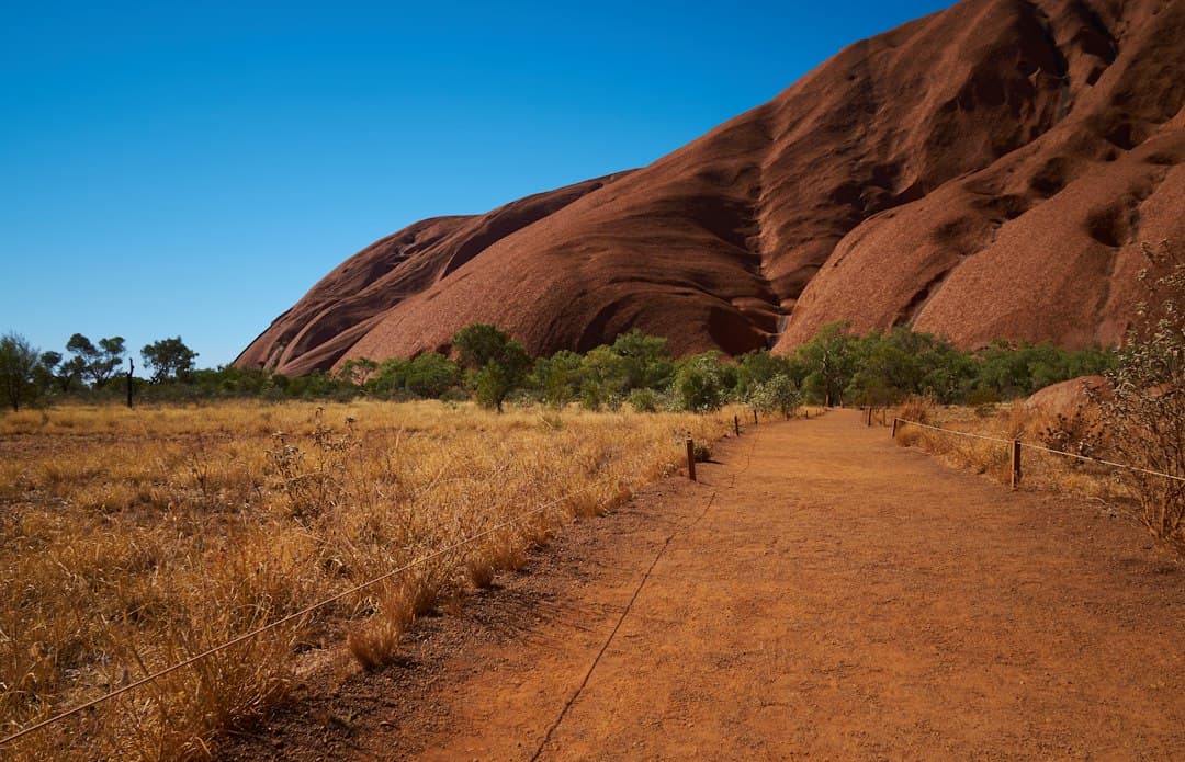 A walking trail at the base of Uluru with rope fencing guidance, showing the iconic red monolith rising against clear sky with sparse desert vegetation on either side. - Photo by Patrick McGregor on Unsplash