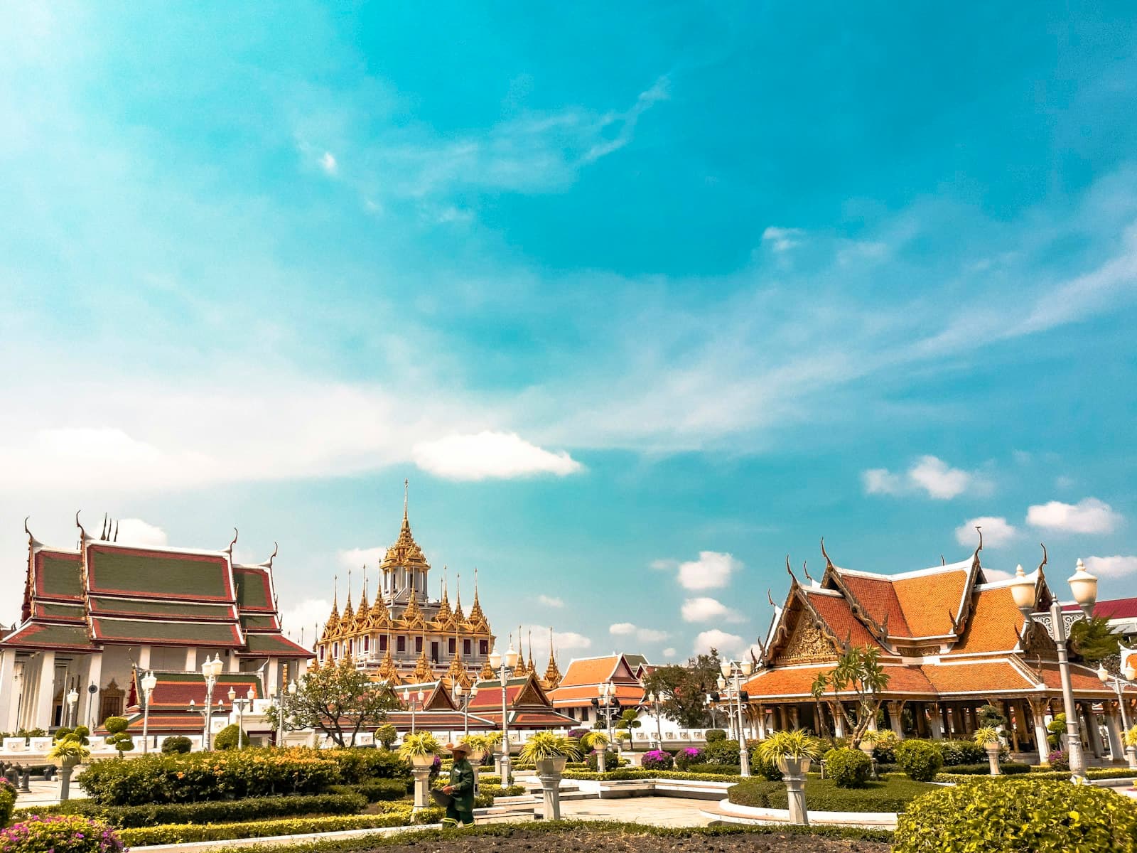 A Thai Buddhist temple complex featuring ornate golden spires, red and gold roofed structures with characteristic upturned eaves, set within manicured tropical gardens under a clear blue sky. - Photo by Alejandro Cartagena 🇲🇽🏳🌈 on Unsplash
