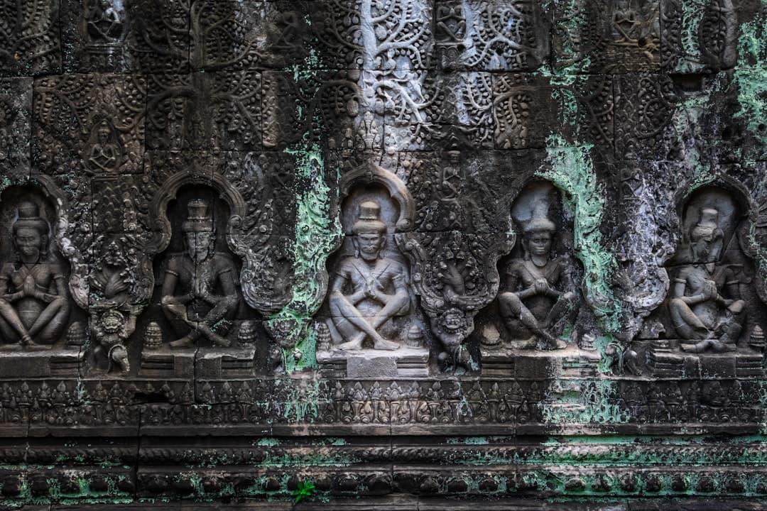 Ornately carved stone relief wall featuring multiple seated figures in meditation poses within arched niches, covered in green lichen and moss, showing weathered Khmer temple architecture. - Photo by Sumit Mangela on Unsplash