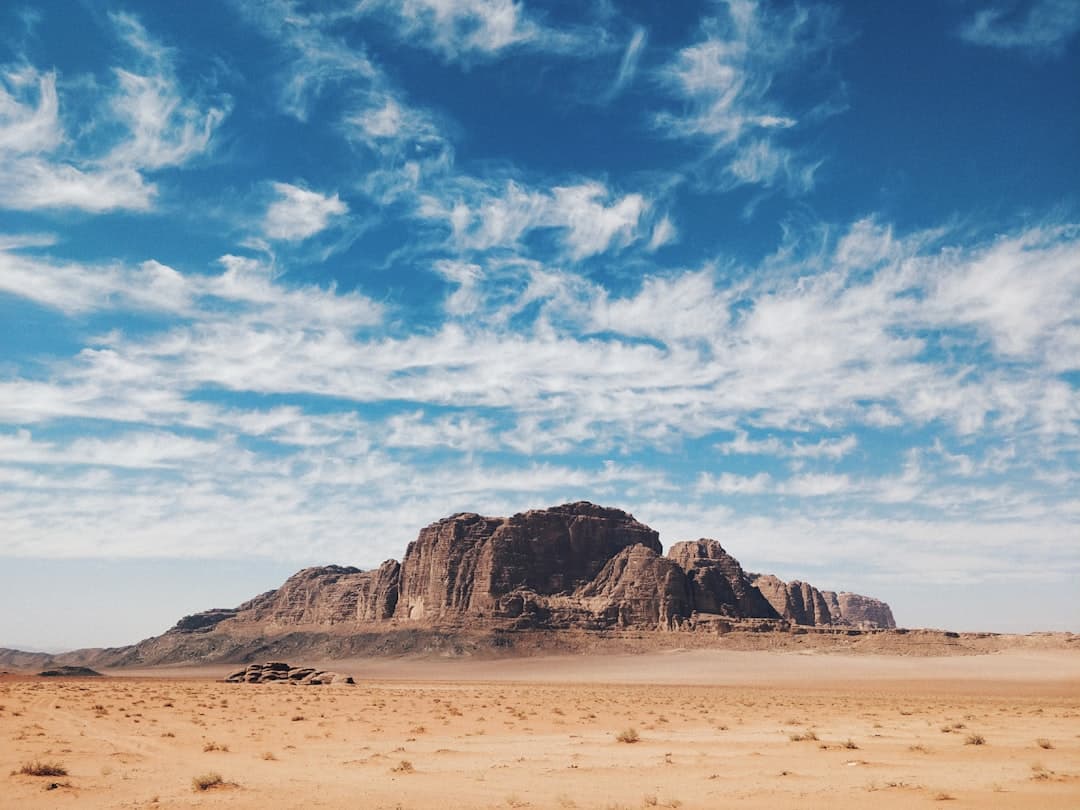 A striking isolated mesa or plateau with vertical columnar rock formations rises from a flat, sparsely vegetated desert plain under a partly cloudy blue sky. - Photo by Anton Lecock on Unsplash
